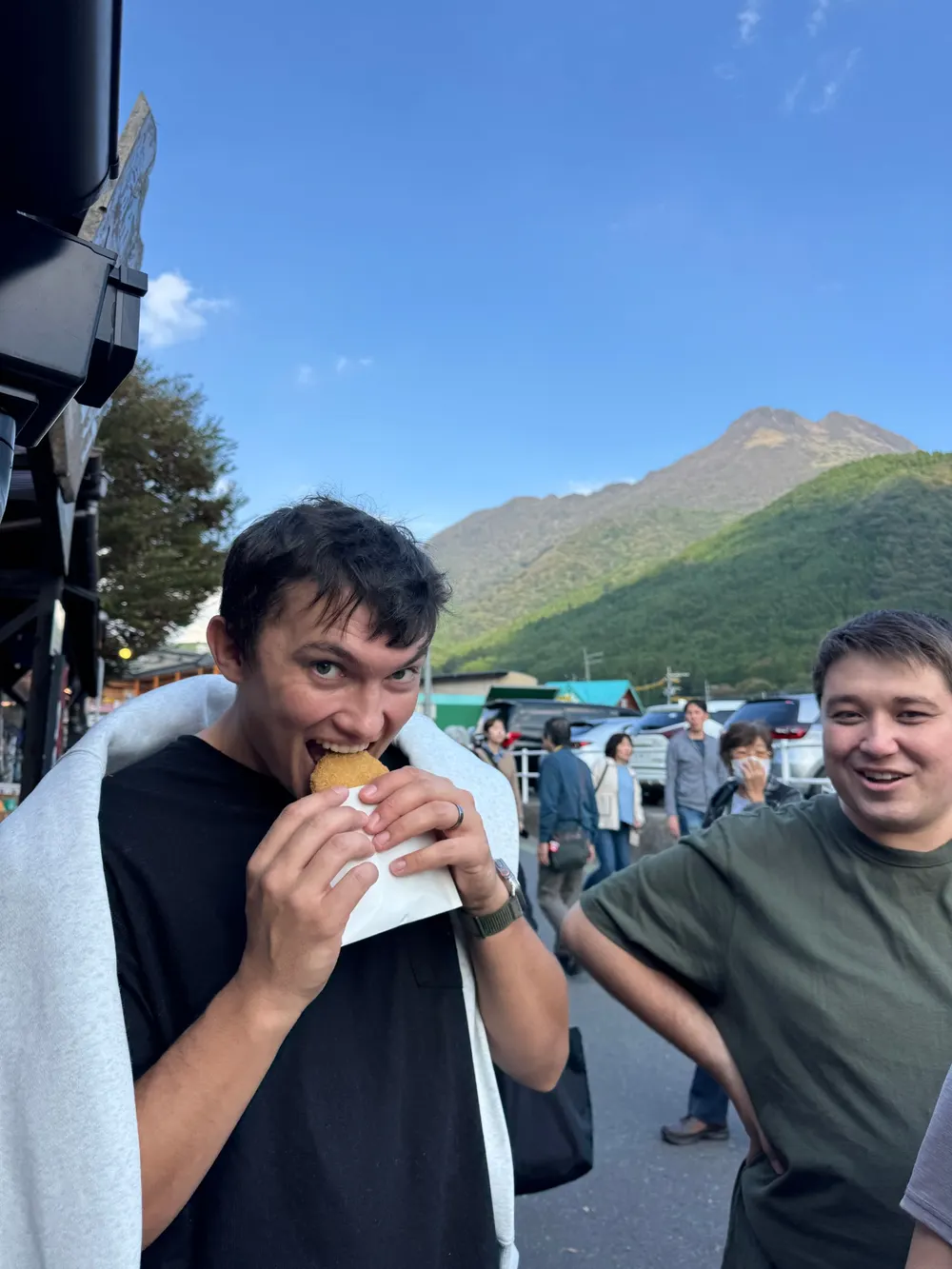 Two travellers eating croquettes on Yufuin's main street, with Yufu-dake behind